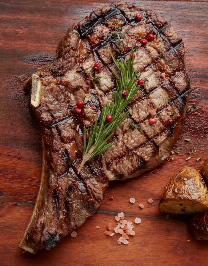 Juicy grilled ribeye steak with rosemary, pink peppercorns, and roasted potatoes on a wooden board.